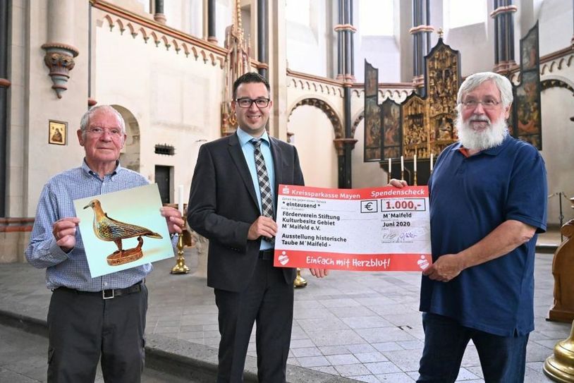 Martin Steinsiek als Leiter des BeratungsCenters der KSK in Münstermaifeld (Mitte) gratuliert den beiden Vorstandsmitgliedern Dieter Ritzenhofen (links) und Otto Wölbert (rechts) persönlich zur Spende und übergibt  symbolisch den „großen“ Scheck im Namen der Kreissparkasse Mayen. Auf dem Bild links ist eine Abbildung der Münstermaifelder Eucharistietaube zu sehen. Foto: KSK