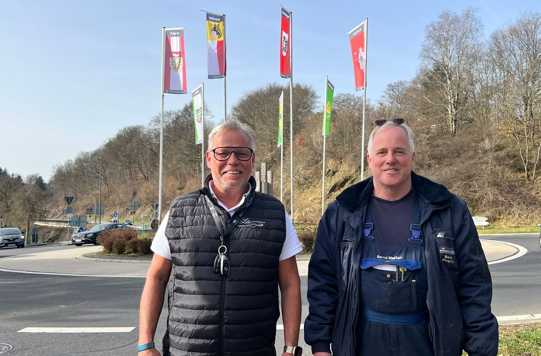 Lutz Schell und Bernd Maaßen (r., beide MONEV) vor den sieben Fahnen der Monschauer Stadtteile, die im Wind auf dem Kreisel an der Monschauer Flora wehen.