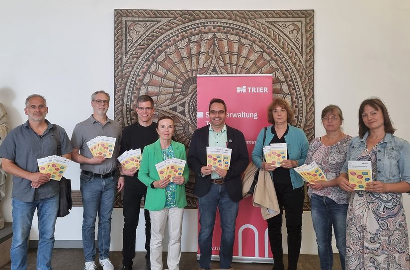 Bürgermeisterin Elvira Garbes (4. v. l.), Sozialraumplaner Simeon Friedrich vom Jugendamt (3. v. l.) und Merit Förster (Bereich Pflegestrukturplanung und Senioren im Sozialamt, r.) präsentieren zusammen mit einigen Vertreterinnen und Vertretern beteiligter Einrichtungen und Träger die neue Auflage der Broschüre: Carsten Schmitt (Treffpunkt am Weidengraben), Andreas Haupenthal (Palais e. V.), Robert Friedrich (Pfarreiengemeinschaft Ehrang-Pfalzel-Biewer), Kerstin Brüning (Fazit e.V. ) und Gabriele Merkel („Gemeinschwester plus“-Team, v. l.)