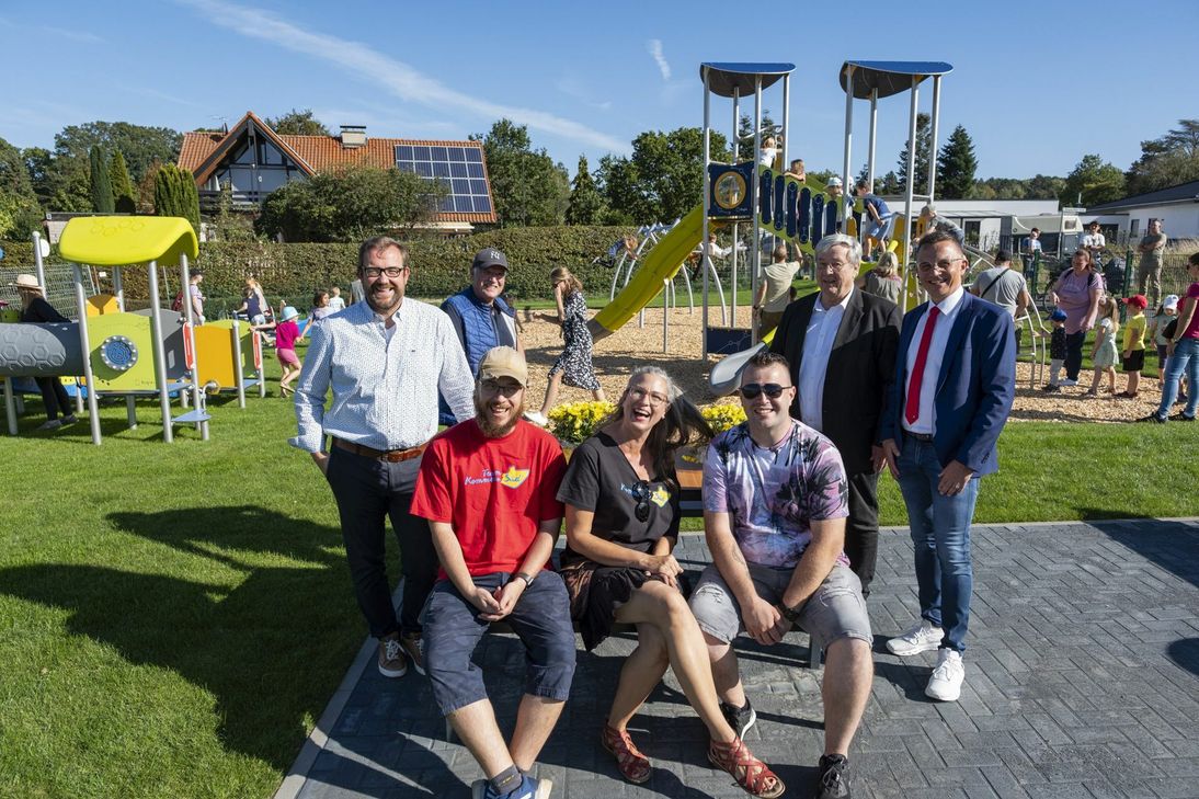 Glückliche Gesichter vorne, spielende Kinder hinten: Zur Spielplatz Eröffnung kamen Mechernichs Beigeordneter Thomas Hambach (stehend, v.l.), Grünflächeningenieur Christof Marx, der stellvertretende Bürgermeister Günter Kornell und Jürgen Hübner als Vertreter des Vorstands der Kreissparkasse. Nathalie Konias wird umrahmt von den Mitstreitern Richard Schlomann (l.) und Tom Ley.