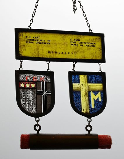 Auf der Hauptplakette finden sich das Kölner Stadtwappen mit den drei Kronen der Heiligen Drei Könige, elf Flammen für die Jungfrauen der heiligen Ursula, das Diözesankreuz, die Buchstaben F, E, K sowie das Papstwappen Johannes Pauls II.