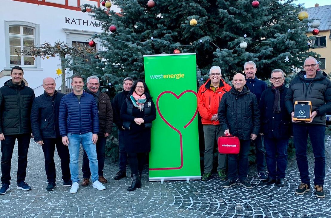 Harald Rosenbaum, Bürgermeister der Verbandsgemeinde Kirchberg, präsentiert gemeinsam mit Florian Schmidt, Regionalmanager bei Westenergie, und einigen der betroffenen Ortsbürgermeister*innen einen von insgesamt zwölf Defibrillatoren.