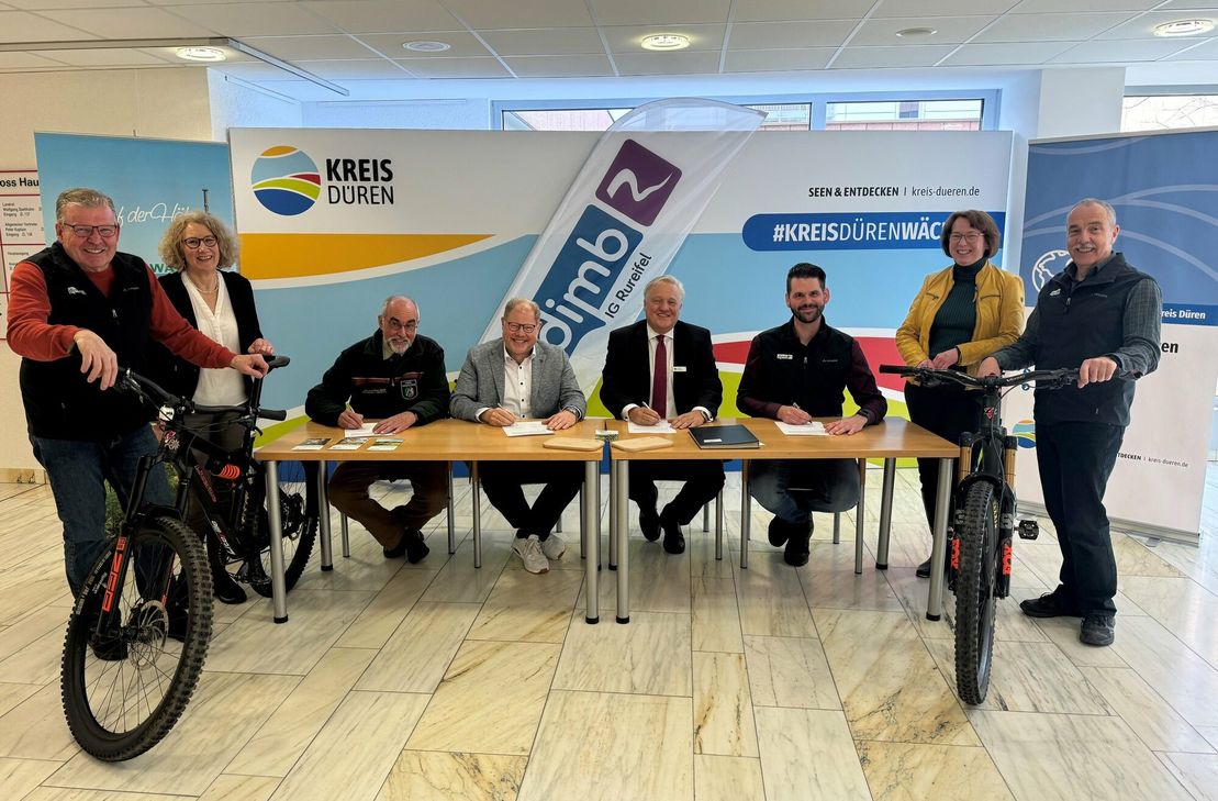 Besiegeln die Zusammenarbeit beim BikePark Rureifel: Robert Jansen (Forstamt), Hürtgenwalds Bürgermeister Stephan Cranen, Landrat Wolfgang Spelthahn, Florian Sporleder (DIMB/v.l. sitzend) im Beisein von Arthur Knobloch (IG Rureifel), Sophia Eckerle, Anette Winkler (beide Kreis Düren) und Axel Volkmer (IG Rureifel/v.l. stehend). Foto: Kreis Düren