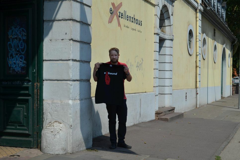 Rino Dzur vom Kulturbüro des Exelllenzhauses mit einem der T-Shirts vor dem Exhaus. Auch Turnbeutel mit dem Logo kann man kaufen. Weitere Artikel sind in Planung. Foto: Urban