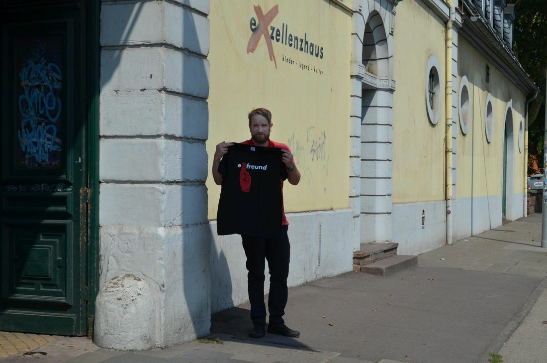 Rino Dzur vom Kulturbüro des Exelllenzhauses mit einem der T-Shirts vor dem Exhaus. Auch Turnbeutel mit dem Logo kann man kaufen. Weitere Artikel sind in Planung. Foto: Urban