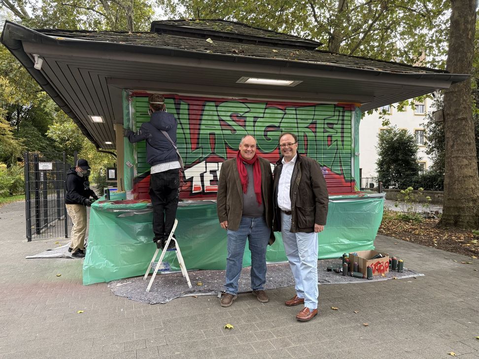 Innenstadtdezernent Ralf Britten und Pächter Niels Becker wirken sichtlich zufrieden vor dem Kiosk im Palastgarten, während die jungen Künstler das neue Graffiti auf den Rollläden gestalten.