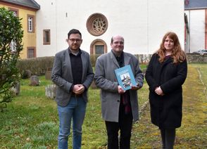 Auf der Kriegsgräberstätte im Schatten der Steinfelder Basilika übergab Andreas Züll (mi.) den Gedenkband zu den Toten des Nationalsozialismus und des zweiten Weltkriegs in der Gemeinde Kall an den Bürgermeister Emmanuel Kunz und Bibliotheksleiterin Michelle Wagner.