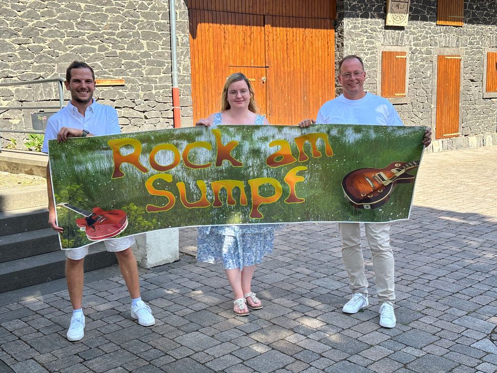 Der Platz an der Thürer Dorfscheune wird am 22. August zum „Rock am Sumpf“-Ground. Ortsbürgermeister Lukas Ellerich (links), Laura Berresheim und Jörg Lempertz, Schirmherr und Bürgermeister VG Mendig, rühren schon einmal die Werbetrommel.