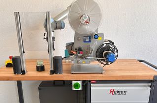 Der Glastiegel (vorne) wird auf dem Arbeitstisch positioniert, Sensoren geben grünes Licht sobald er an der richtigen Stelle ist. Dann klebt der Cobot das Etikett darauf.