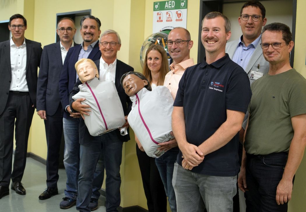 Gruppenbild mit Übungspuppen und AED:  André Polrolniczak und Martin Grünen (Vorstandsmitglieder Sparkasse Trier), Ivan Racic (Geschäftsführer Nikolaus Koch Stiftung), Manfred Bitter (Vorstandsvorsitzender Nikolaus Koch Stiftung und Vorstandsmitglied Herbert &amp; Veronika Reh Stiftung), Carsten Stiller und Amelie Schäfer (Humboldt-Gymnasium Trier) sowie Tobias Hauptmann (Hand aufs Herz Trier e.V.), Michael Molitor (stellvertretender Hausoberer Brüderkrankenhaus Trier) und Dr. med. Thomas Gehrig (Hand aufs Herz Trier e.V.)