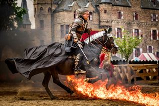 Ein Ritter auf Pferd reitet durch ein spektakuläres Feuerspiel – im Hintergrund die mittelalterliche Burg Satzvey.
