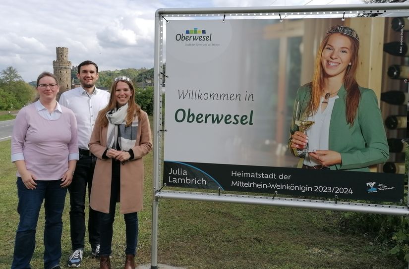 Zwei große Banner schmücken den Straßenrand: Mittelrhein-Weinkönigin Julia Lambrich (rechts) stellt gemeinsam mit Stadtbürgermeister Marius Stiehl (Mitte) und Franziska Schallop, Leitung der Tourist-Information (links) die neuen Banner vor.