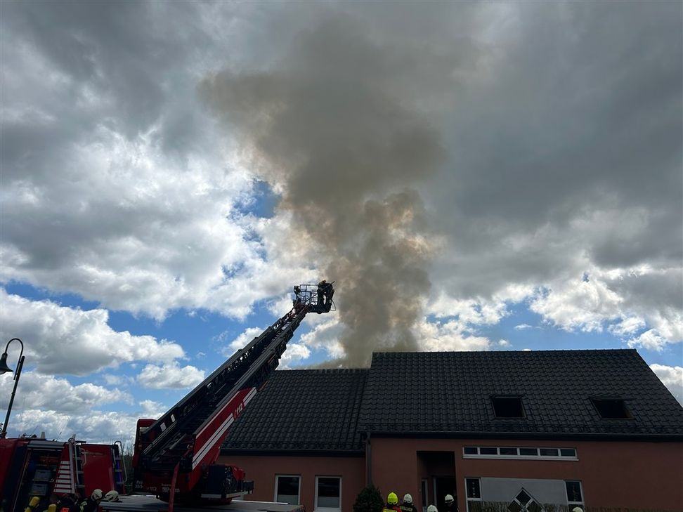 Ein Feuerwehreinsatz bei einem Hausbrand unter bewölktem Himmel. Eine rot-weiße Drehleiter ist weit ausgefahren; im Korb an der Spitze befinden sich Feuerwehrleute, die gegen eine dichte, gräuliche Rauchwolke vorgehen, die aus dem Dachstuhl eines zweistöc