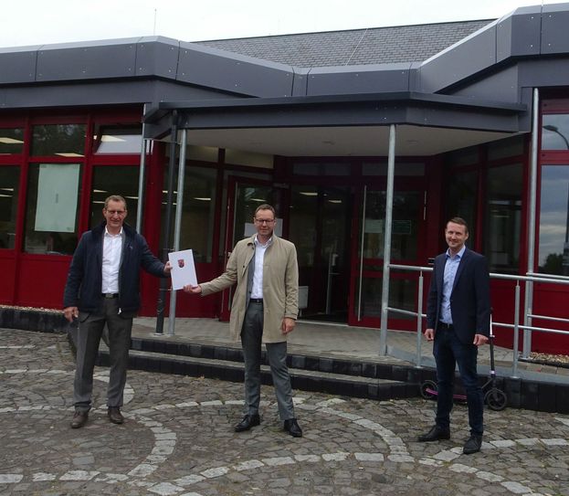 Bürgermeister Maximilian Mumm und Thomas Häuser (rechts) vom Bauamt der VG Maifeld freuen sich über den von Staatssekretär Dr. Alexander Wilhelm überreichten Förderbescheid für die Maifeldhalle in Polch. Foto: VG Maifeld