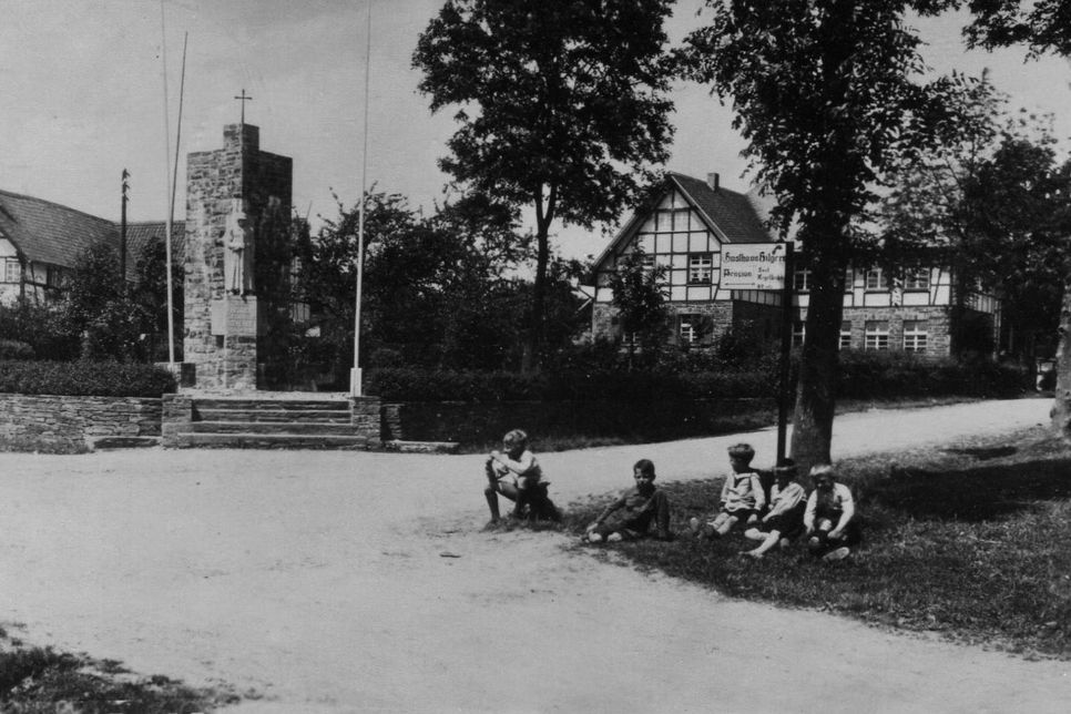 Kinder im Vorgarten des heutigen Autohauses Offermann in Eicherscheid mit Kriegerdenkmal und Kindergarten im Hintergrund. Foto: AK Geschichte Eicherscheid