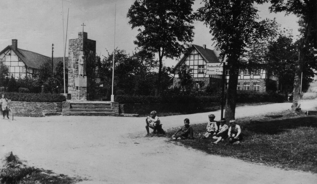 Kinder im Vorgarten des heutigen Autohauses Offermann in Eicherscheid mit Kriegerdenkmal und Kindergarten im Hintergrund. Foto: AK Geschichte Eicherscheid