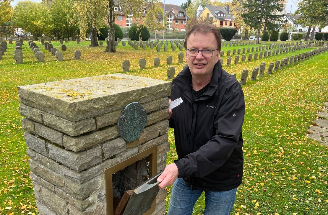 Diakon Jens Schramm öffnet die Klappe der Stele auf dem Ehrenfriedhof in der ein Namensverzeichnis der dort beigesetzten 563 Kriegstoten untergebracht werden soll.  Zudem werden QR-Codes etwas über  die Geschichte einiger der Beigesetzten erzählen.