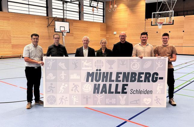 Sie halten das neue Schild der Mühlenberghalle (v.l.): Armin Fischbach (Fachbereichsleiter Hochbau), Marcus Bierschenk (Büro abph Heimbach), Wolfgang Schmitz (Astrid-Lindgren-Schule), Lene Drude (Grundschule Schleiden), Bürgermeister Ingo Pfennings, Felix Stoff (Stadt Schleiden, Hochbau) und Sven Löhr (Grundschule Schleiden).
