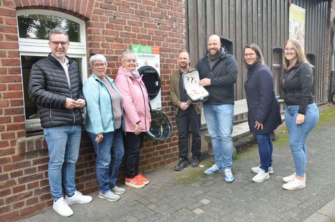 Bürgermeister Norbert Crump (v.li.), Angela Mauel und Elfriede Schneider, Basargemeinschaft Nettersheim, Dennis Matull, Geschäftsführer ALDI Zingsheim, Michael Gissinger, Verein Lebensretter, sowie Eva Gäbler und Isabella Mauel, Eifelgemeinde Nettersheim, stellen den Defibrillator an der Alten Schmiede vor.